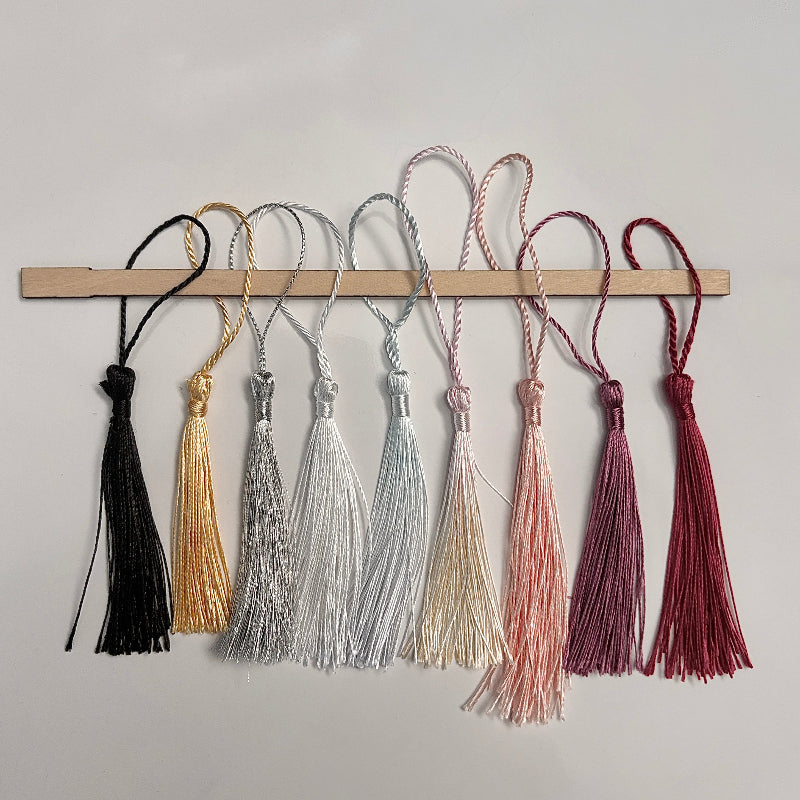Set of six tassels in black, gold, silver, beige, pink, and red on a wooden rod against a gray background.